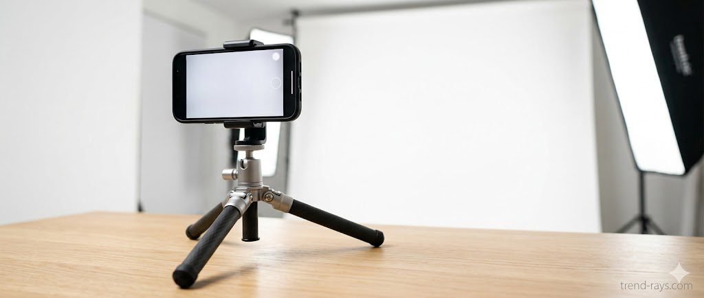 A high-quality, modern header graphic showing an iPhone mounted on a sleek desktop tripod, angled towards a clean white backdrop. Soft, professional lighting illuminates the scene.